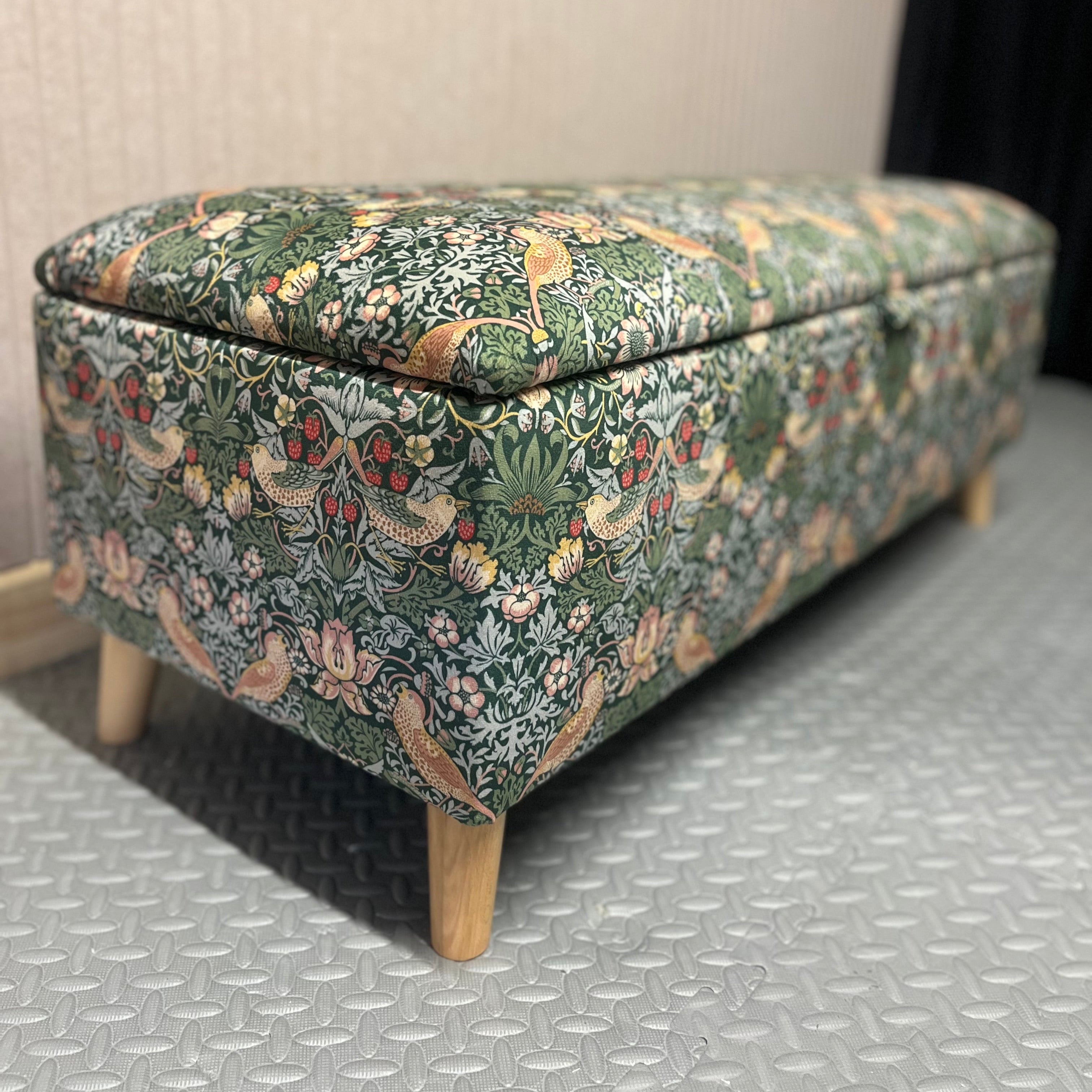 FLoral-pattern ottoman in a velvet William morris print with a green background. Wooden angled legs on a textured floor