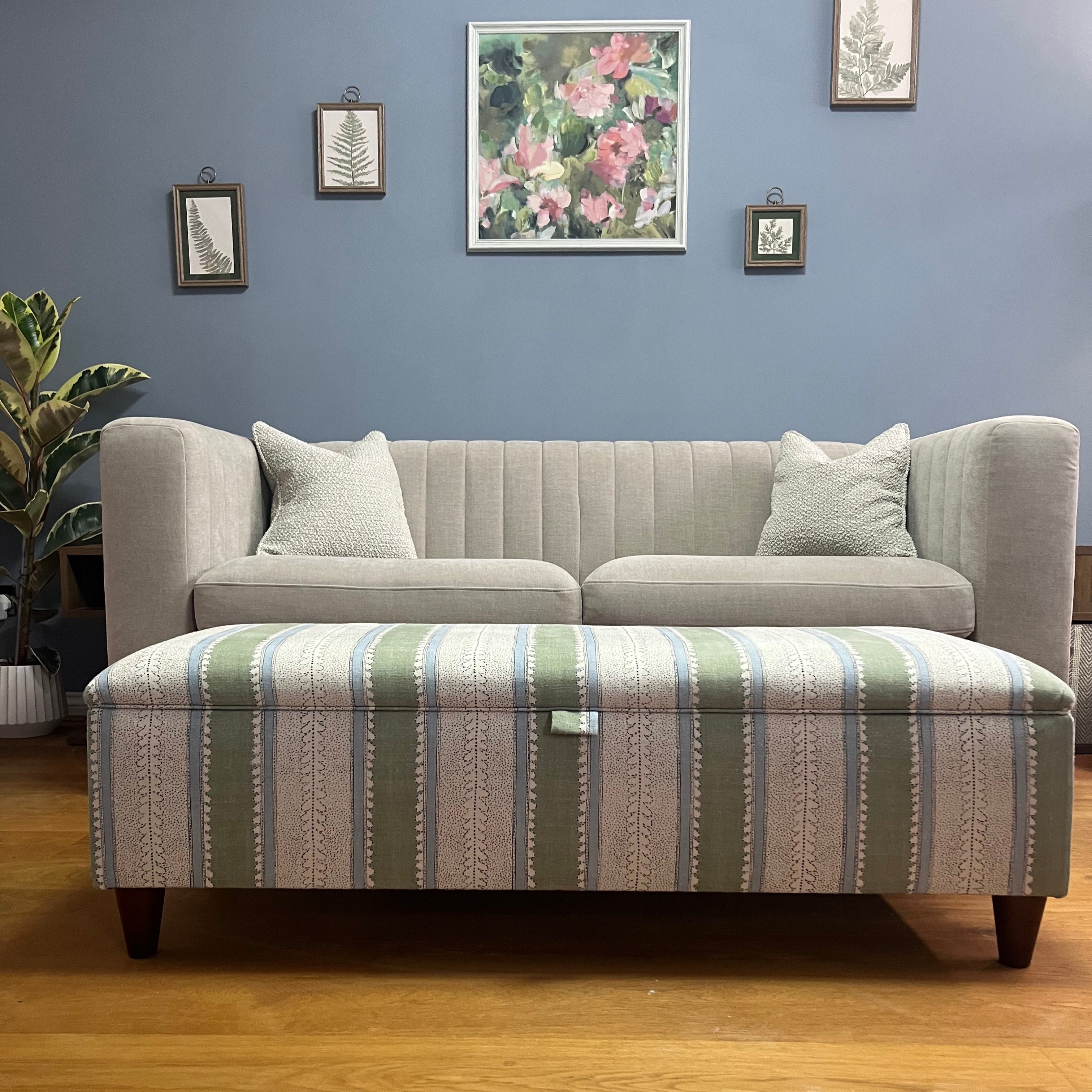Grey upholstered sofa with striped storage ottoman against a blue wall with framed pictures