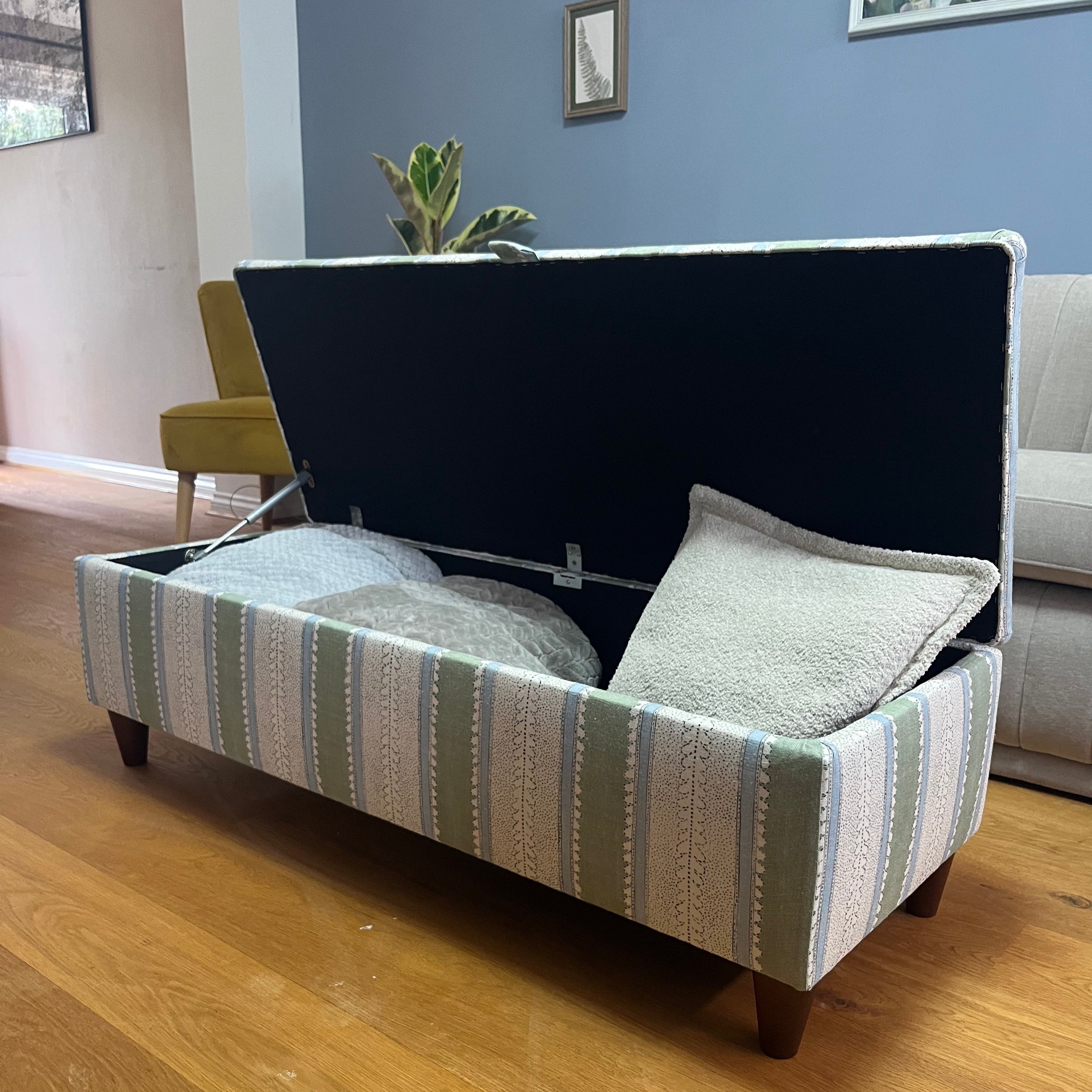 Green, blue and white striped ottoman with walnut legs, open with blankets and cushions inside.