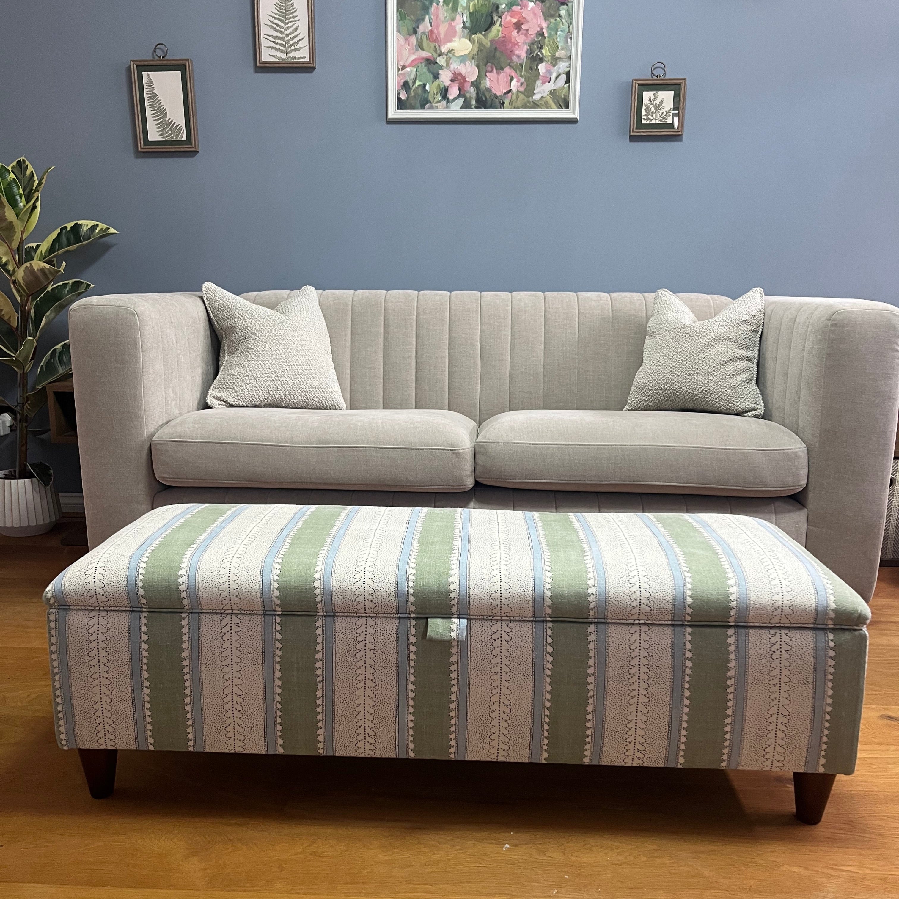 Grey sectional sofa with stripes storage footstool against a blue wall with framed pictures.