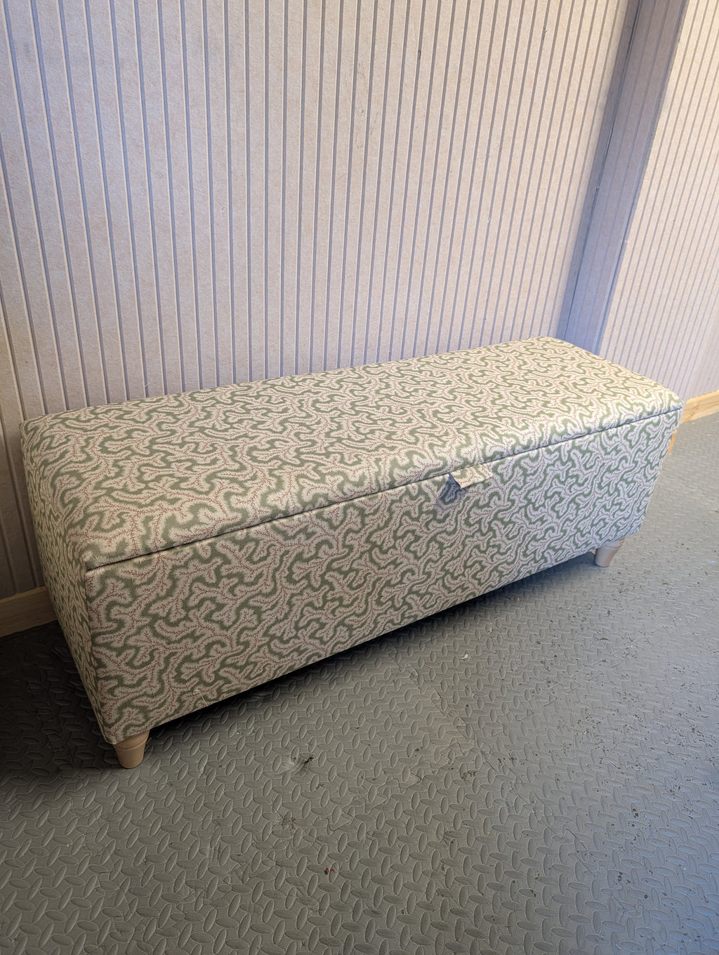 An upholstered storage bench with a geometric patterned fabric in  pink and green, set against a light-colored paneled wall on a gray carpeted floor.