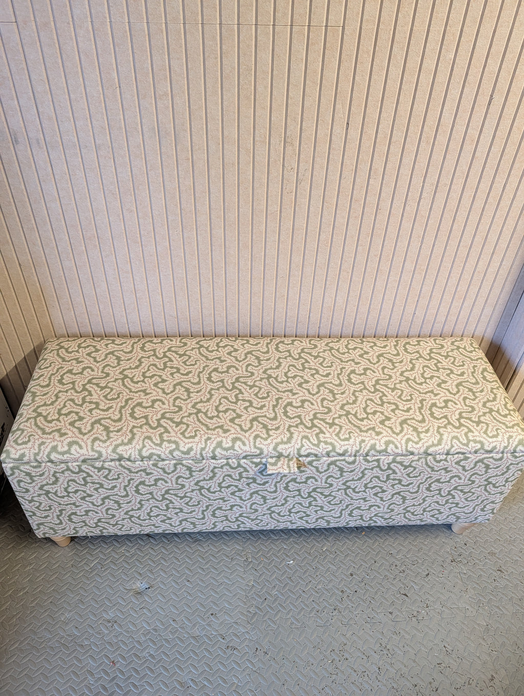 An upholstered storage bench with a light green and pink wavy patterned fabric, set against a beige ribbed wall on a gray textured floor.