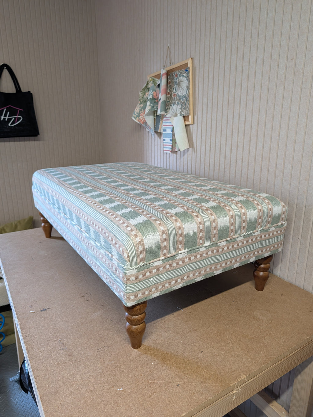 An upholstered ottoman or bench with a light blue and beige geometric pattern, supported by four turned wooden legs, shown on a workbench in a room with striped wallpaper.