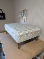 An upholstered ottoman or bench with a light blue and beige geometric pattern, supported by four turned wooden legs, shown on a workbench in a room with striped wallpaper.