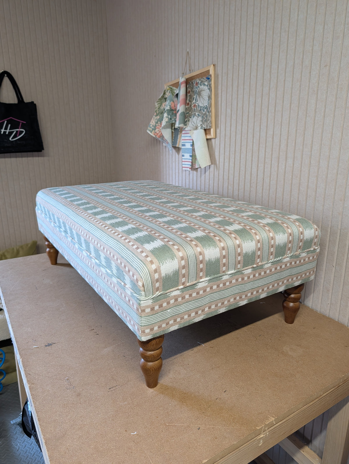 An upholstered ottoman or bench with a light blue and beige geometric pattern, supported by four turned wooden legs, shown on a workbench in a room with striped wallpaper.