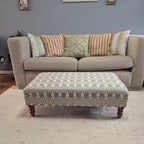 An upholstered rectangular ottoman with a geometric pattern in sage green, beige, and white, supported by four turned wooden legs, positioned in front of a light beige sofa with decorative throw pillows on a cream shag rug.