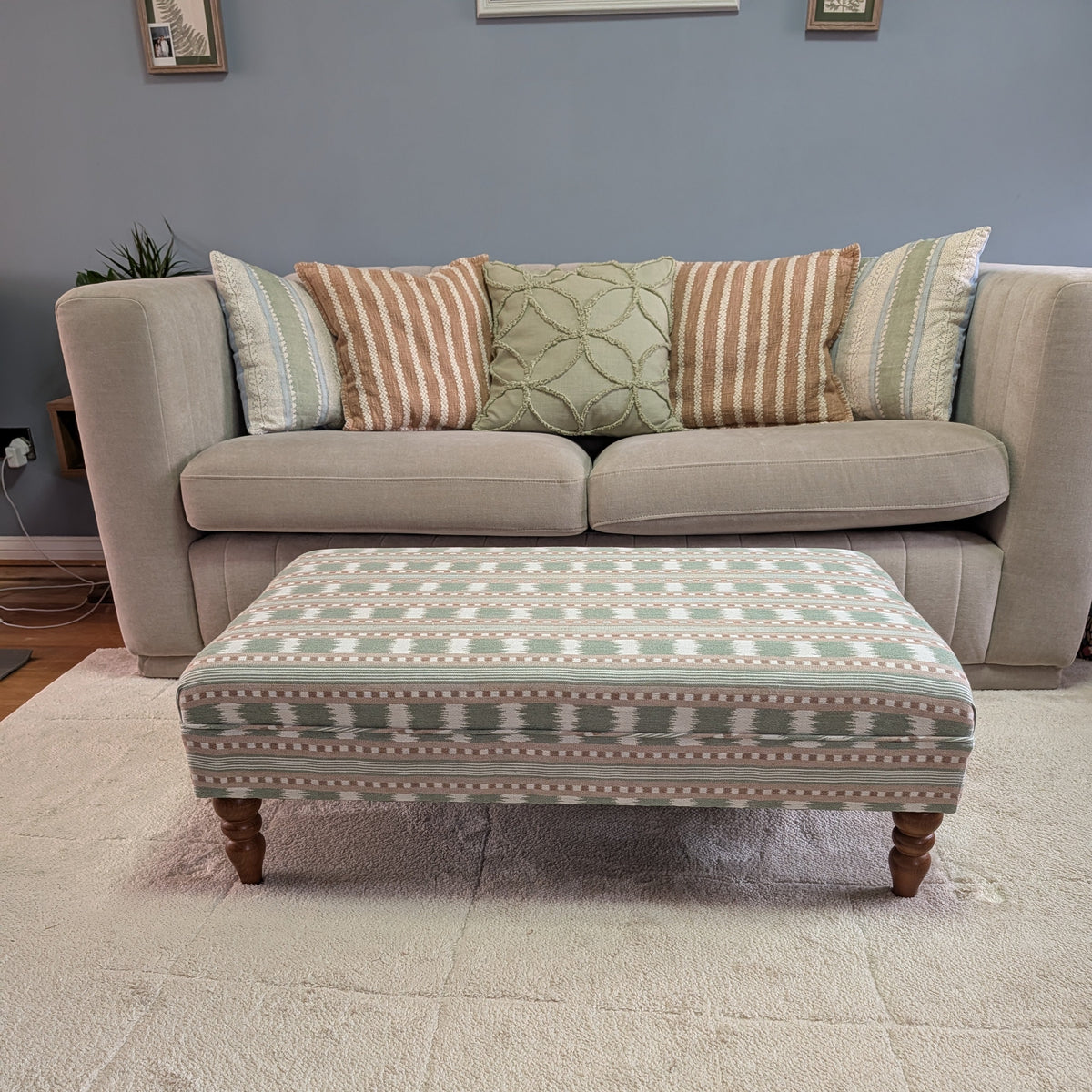 An upholstered rectangular ottoman with a geometric pattern in sage green, beige, and white, supported by four turned wooden legs, positioned in front of a light beige sofa with decorative throw pillows on a cream shag rug.