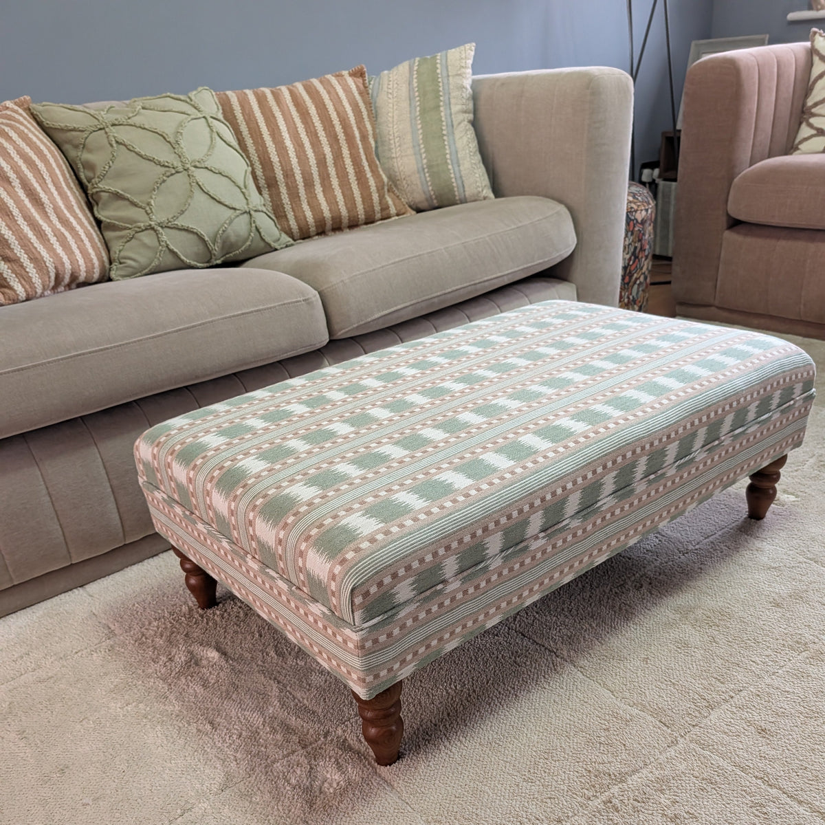 An upholstered rectangular ottoman with a geometric pattern in sage green, pink, and beige, supported by dark wooden legs, shown in a living room setting on a beige rug in front of a sofa.
