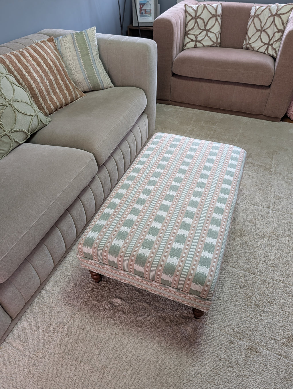 A rectangular upholstered ottoman with a pink, green, and beige geometric pattern sits on a light beige rug in a living room next to a gray sofa.