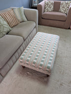 A rectangular upholstered ottoman with a pink, green, and beige geometric pattern sits on a light beige rug in a living room next to a gray sofa.