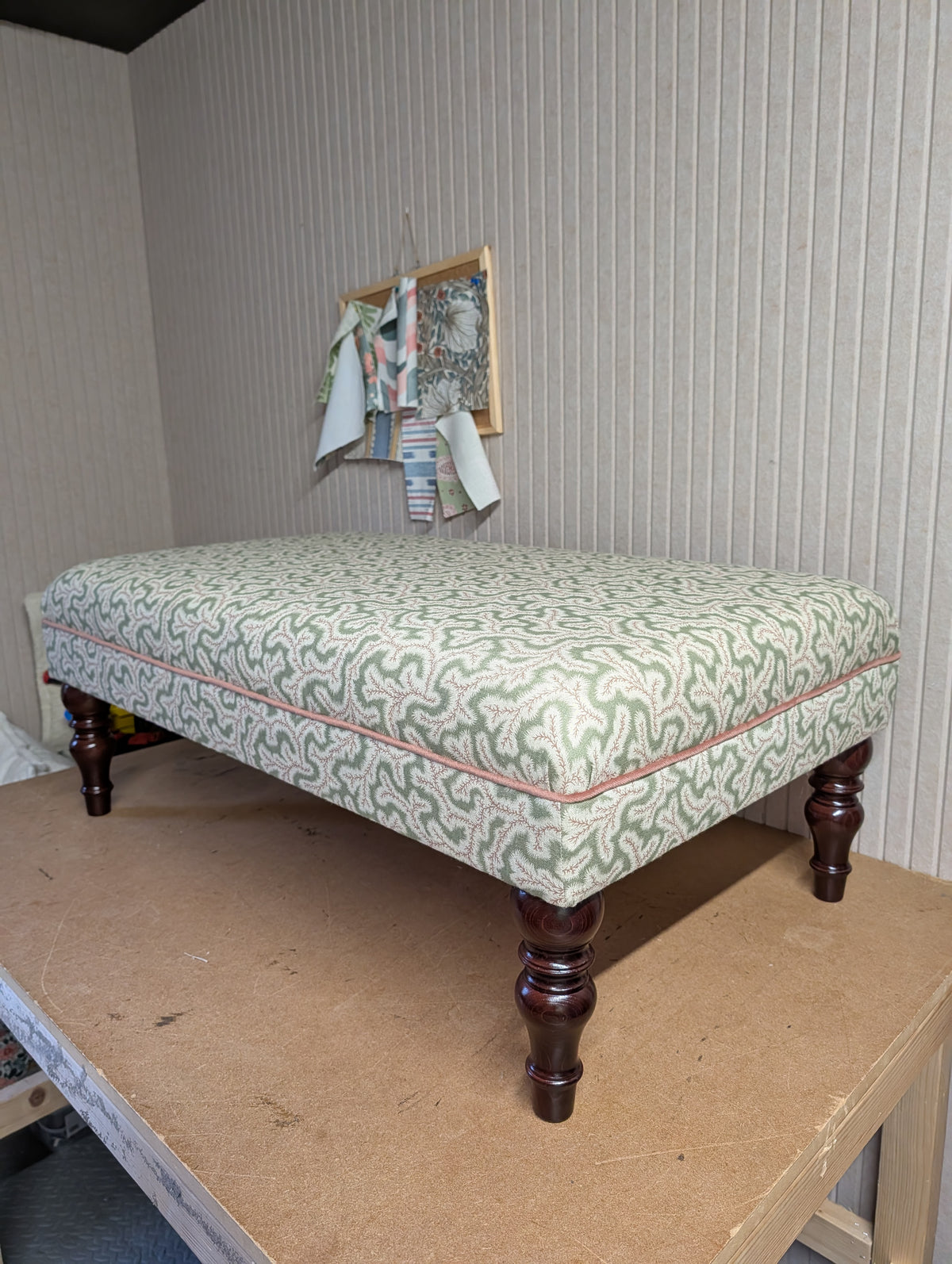 An upholstered bench or ottoman with a green and white wavy patterned fabric, dark brown turned wooden legs, and a thin pink piping trim, shown on a workbench in a room with ribbed wallpaper.
