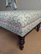 An upholstered bench or ottoman with a green and white coral-like patterned fabric, pink piping along the edge, and dark brown turned wooden legs, shown in a room setting.