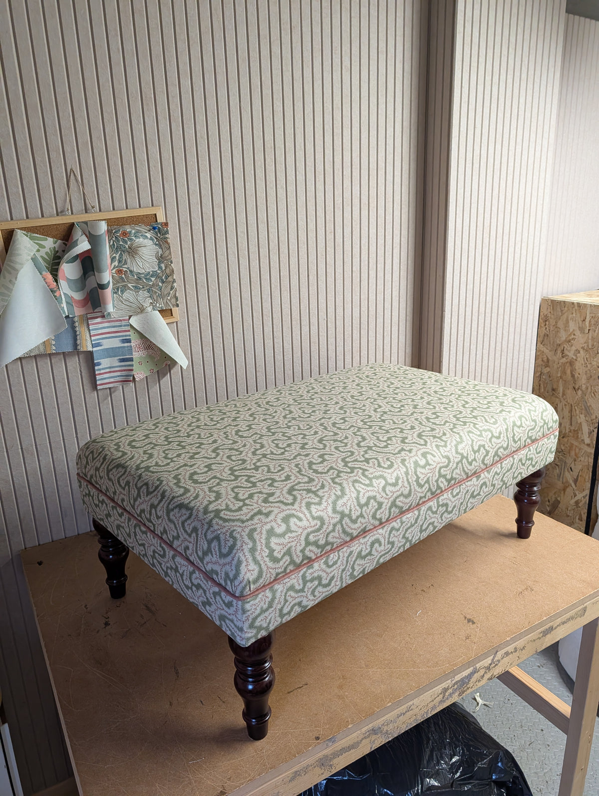 An upholstered rectangular ottoman with a green and beige wavy patterned fabric, dark brown turned legs, and a thin rose pink piping detail along the front edge, shown on a wooden workbench in a workshop setting.