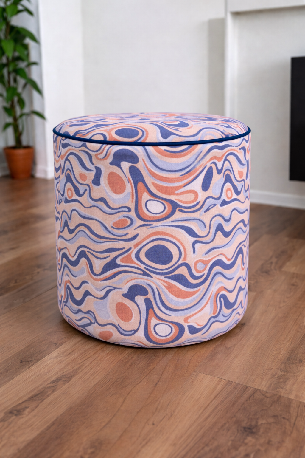 A cylindrical upholstered ottoman with a swirling abstract 70s pattern in navy blue, peach, and off-white, set on a wooden floor in a bright room.