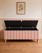 A striped fabric storage bench with an open lid, featuring red and cream vertical stripes, light wood legs, and a black fabric interior, set against a cream wall with a framed botanical print and light wood floor.