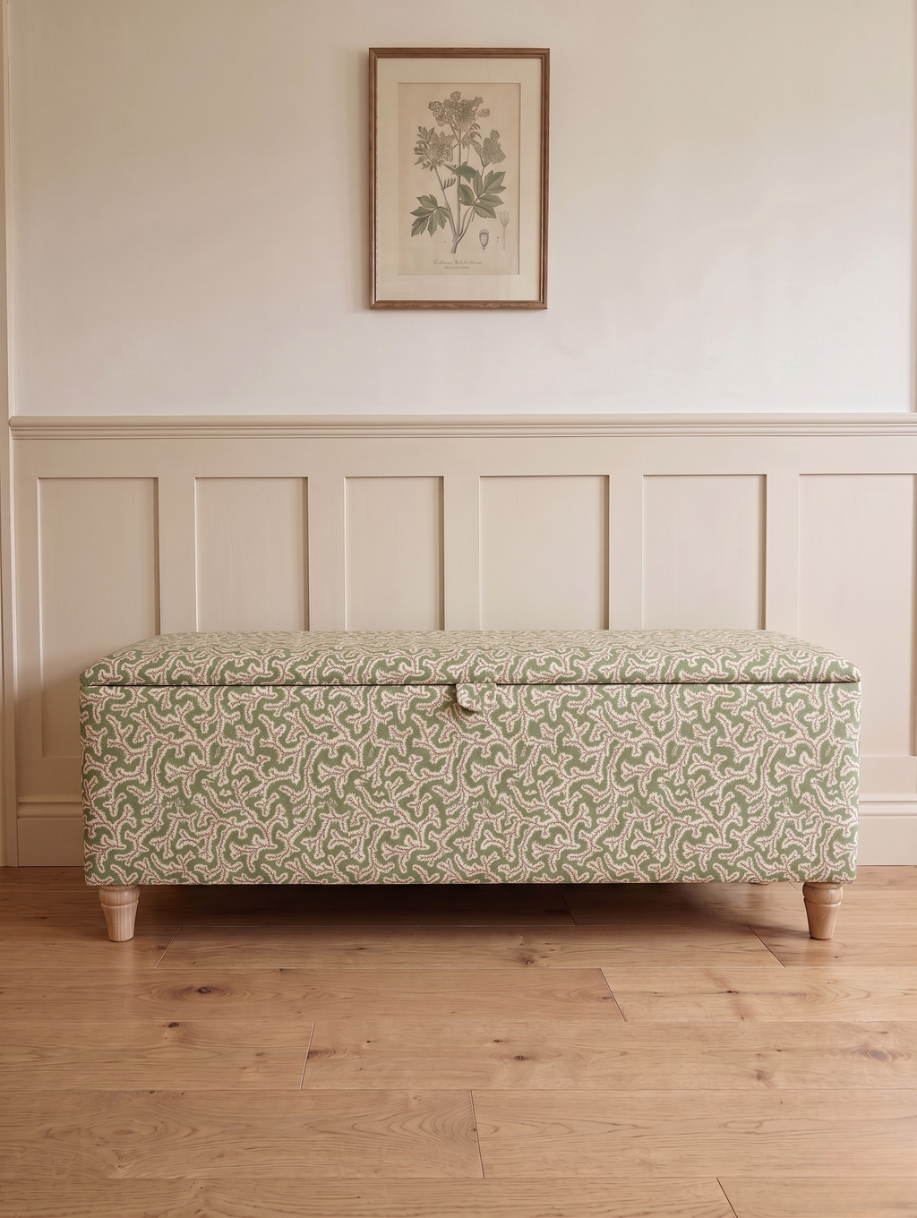 A storage bench with a light green and pink floral patterned Ian Sanderson dulcie fabric upholstery, standing on a light wood floor against a cream paneled wall with a framed botanical print.