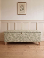 A storage bench with a light green and pink floral patterned Ian Sanderson dulcie fabric upholstery, standing on a light wood floor against a cream paneled wall with a framed botanical print.