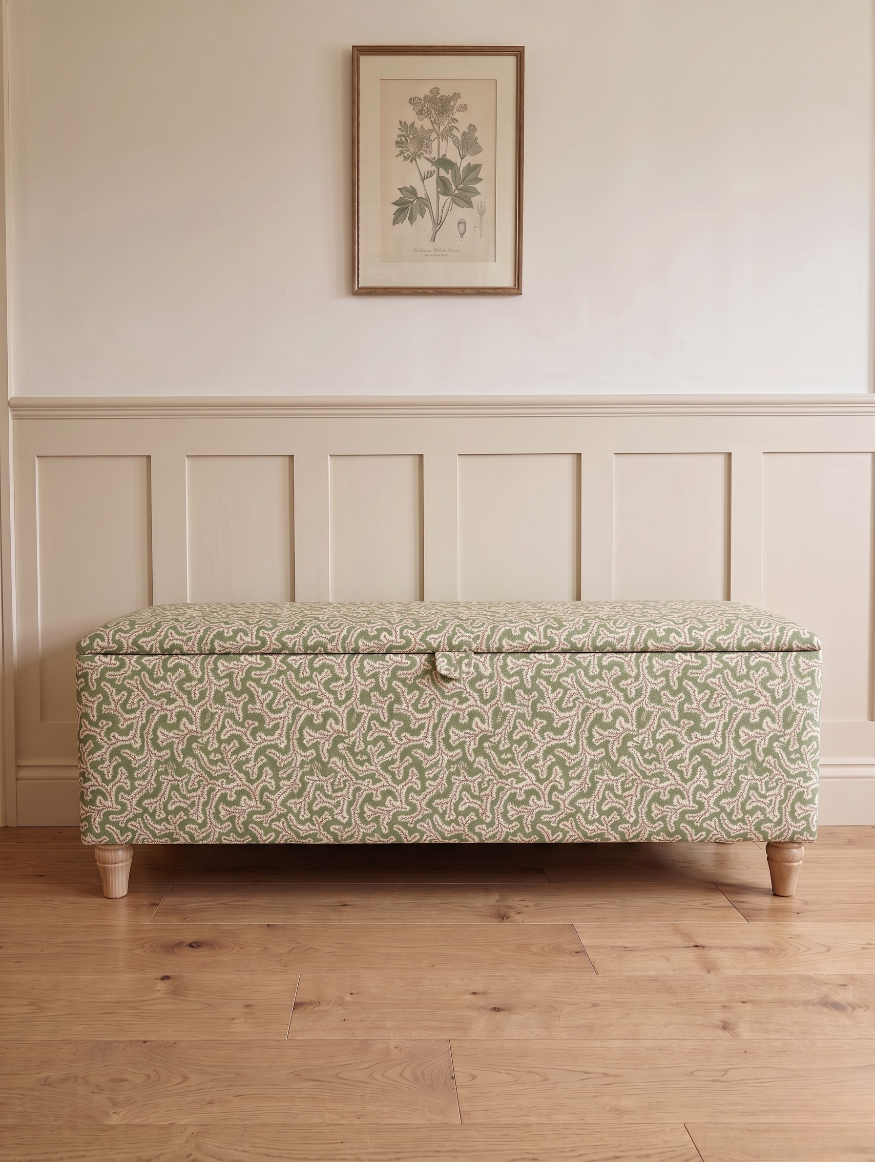 A storage bench with a light green and pink floral patterned Ian Sanderson dulcie fabric upholstery, standing on a light wood floor against a cream paneled wall with a framed botanical print.