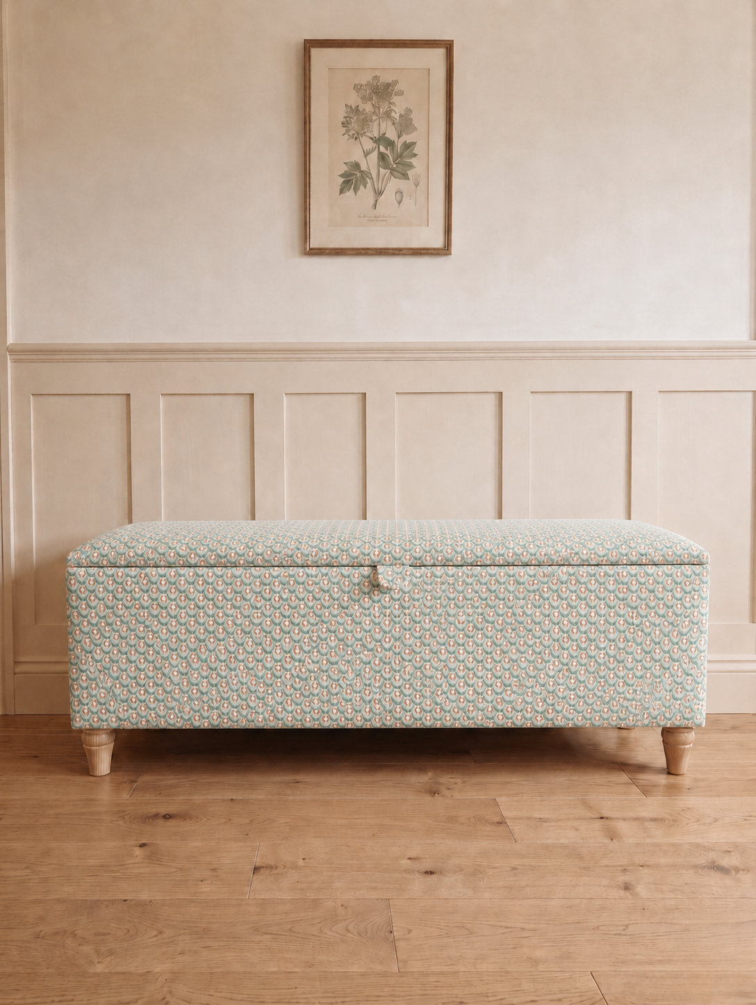 A storage bench upholstered in a pale sage green and white scallop patterned fabric, set against a cream panelled wall and light wood floor.