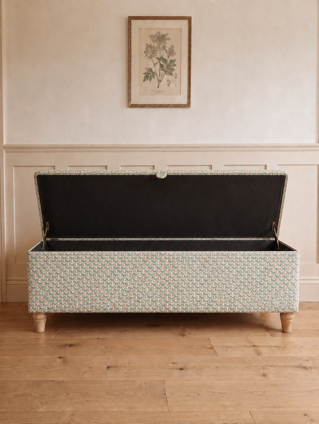 A storage bench with a light beige and white scallop geometric patterned upholstery, shown open to reveal a black fabric-lined interior. The bench is positioned on a light wood floor against a cream-colored wall with decorative paneling and a framed botanical print.