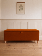 A rectangular upholstered storage bench in a burnt orange amber fabric with turned wooden legs, set against a cream panelled wall with a framed botanical print above.