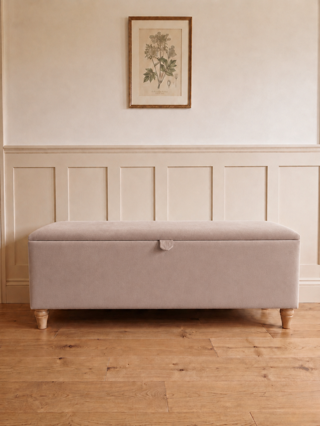 A rectangular upholstered storage bench with a light beige fabric cover and turned wooden legs, placed in a room with wood flooring, white paneling, and a framed botanical print on the wall.
