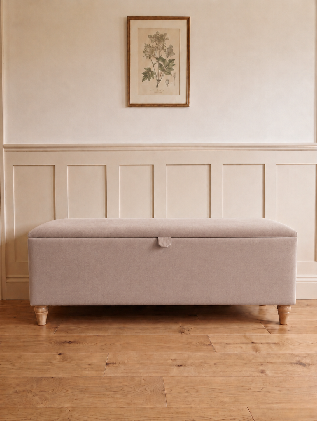 A rectangular upholstered storage bench with a light beige fabric cover and turned wooden legs, placed in a room with wood flooring, white paneling, and a framed botanical print on the wall.
