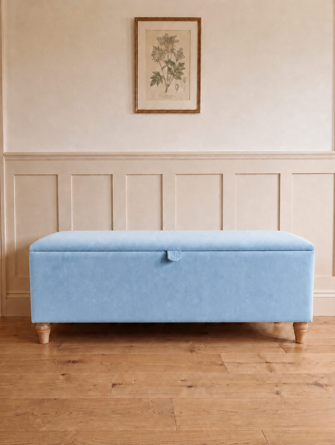 A light blue upholstered storage bench with wooden legs, set against a cream paneled wall with a framed botanical print above.