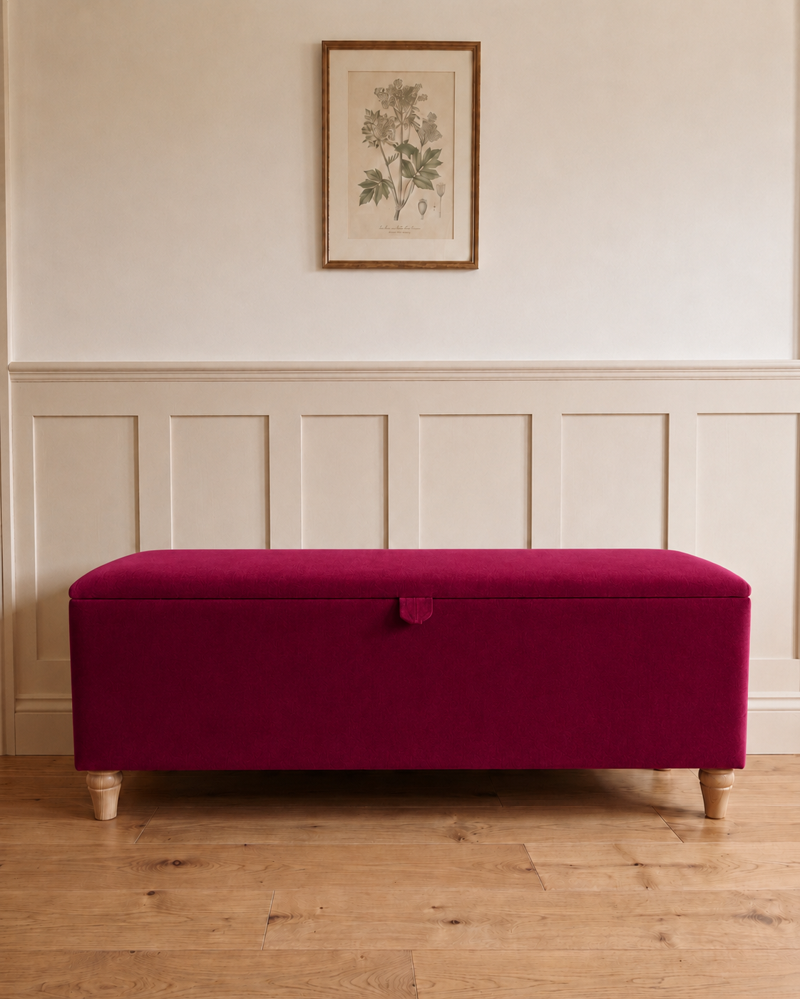 A rectangular upholstered storage bench in rich magenta raspberry velvet with light wood legs, set against a white paneled wall with a framed botanical print above.