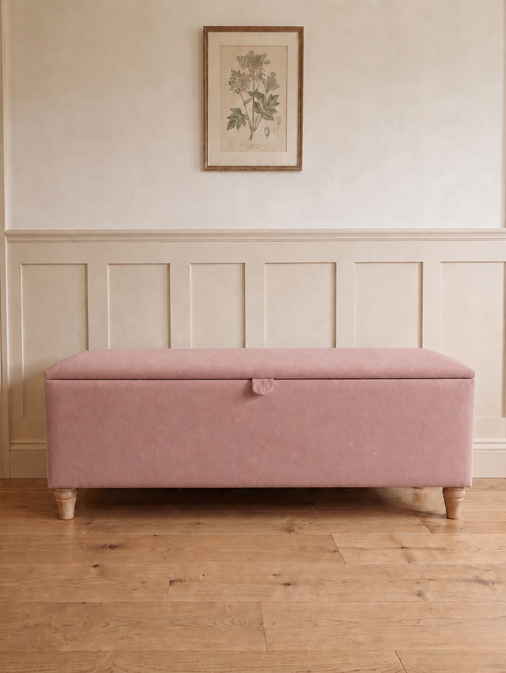 A rectangular upholstered storage bench in a rose pink green fabric with turned wooden legs, set against a cream panelled wall with a framed botanical print above.