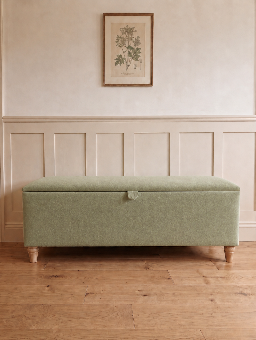A rectangular upholstered storage bench in a muted sage green fabric with turned wooden legs, set against a cream paneled wall with a framed botanical print above.