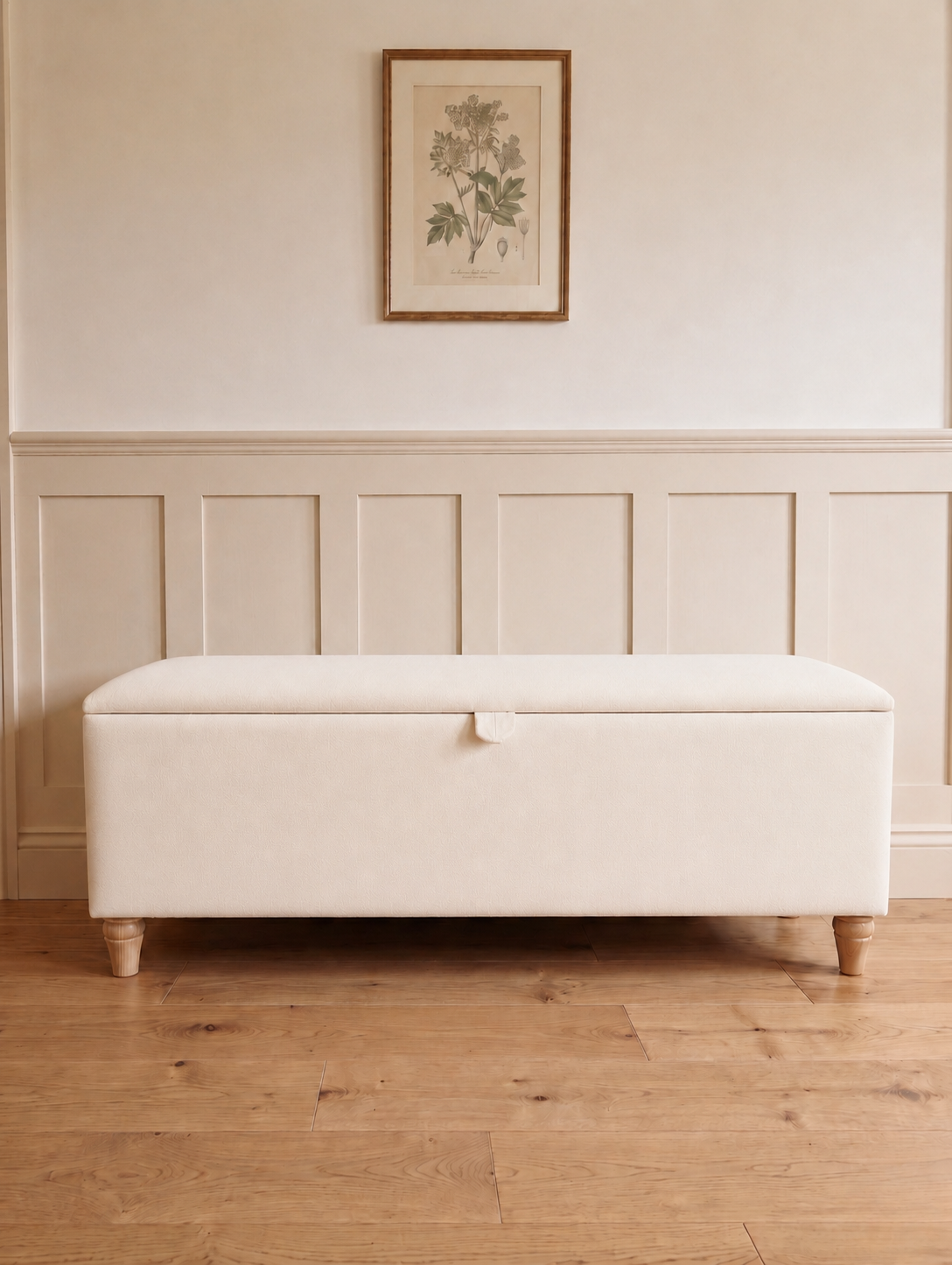 An upholstered storage bench with a hinged lid, resting on a light wood floor against a cream paneled wall with a framed botanical print.