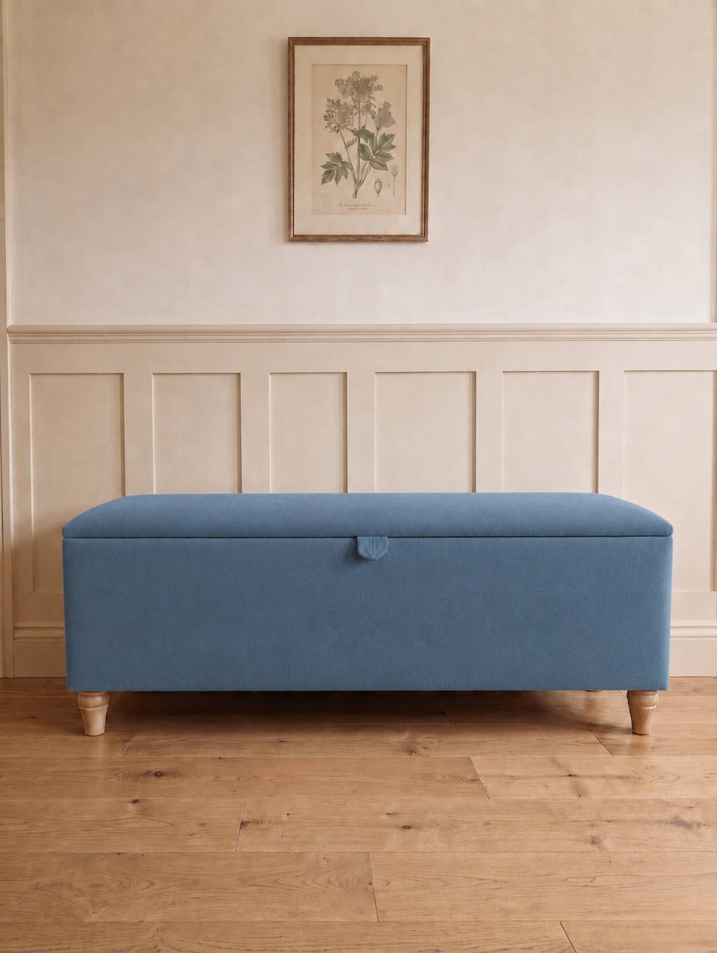 A blue upholstered storage bench with wooden legs, set against a cream paneled wall with a framed botanical print above.