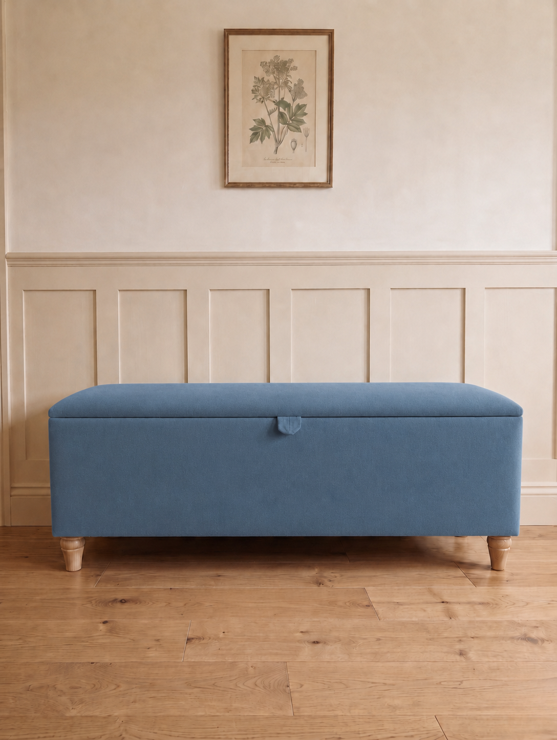 A blue upholstered storage bench with wooden legs, set against a cream paneled wall with a framed botanical print above.
