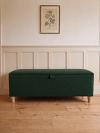A dark green upholstered storage bench with wooden legs, set against a white paneled wall with a framed botanical print above.