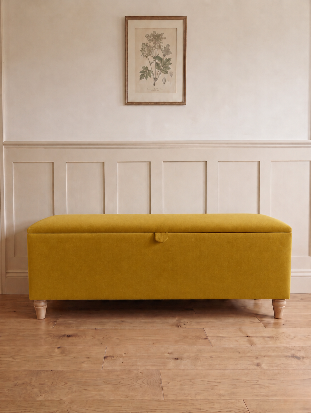 A rectangular upholstered storage bench in a honey yellow fabric with turned wooden legs, set against a cream panelled wall with a framed botanical print above.