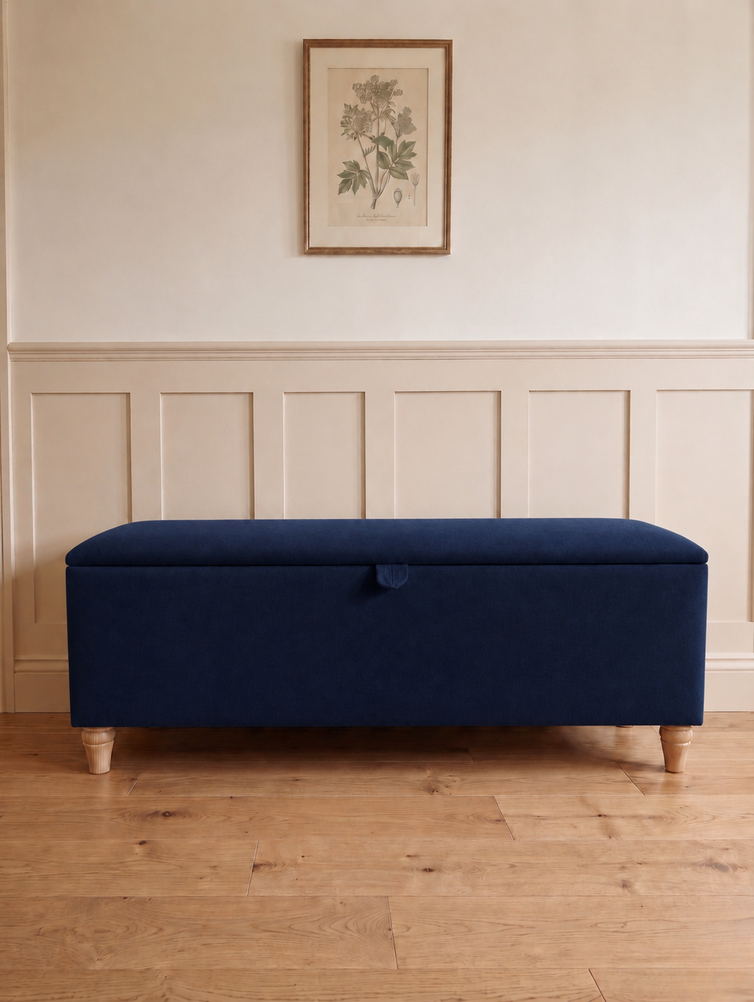 A rectangular upholstered storage bench in a royal blue fabric with turned wooden legs, set against a cream panelled wall with a framed botanical print above.