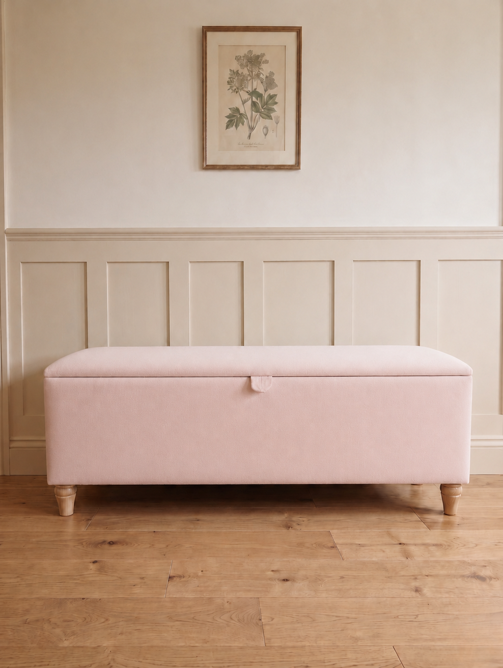 A light pink upholstered storage bench with wooden legs, set against a cream paneled wall with a framed botanical print above.