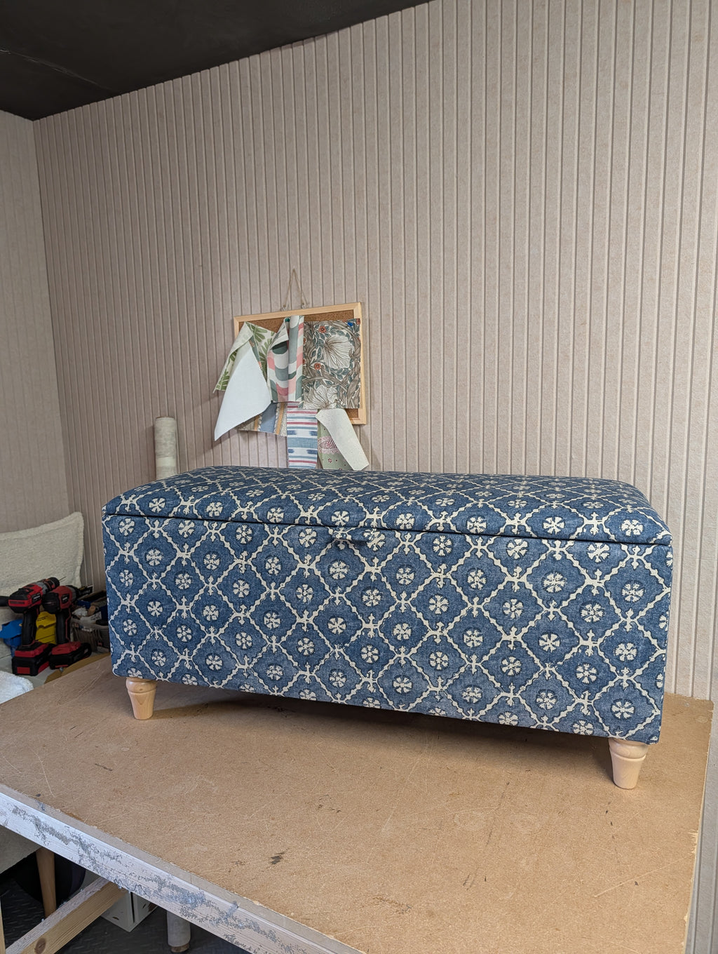 A rectangular storage bench with a blue and white geometric patterned fabric upholstery and light wood legs, set on a wooden workbench against a beige ribbed wall.