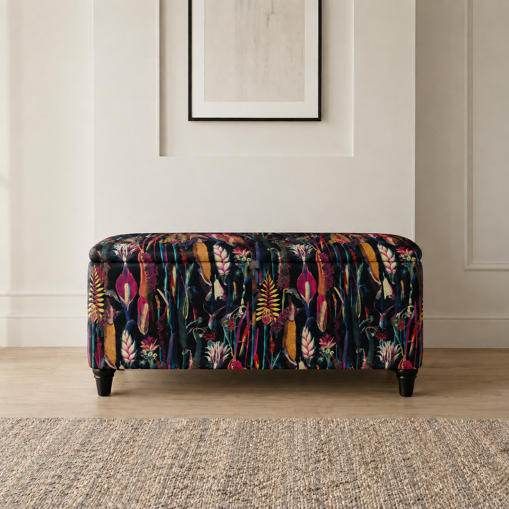 A rectangular upholstered bench with a colorful botanical and floral pattern on a dark background, supported by four black legs, set in a room with a light wood floor and a textured rug.