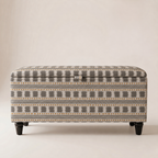 An upholstered storage ottoman with a geometric pattern in gray, beige, and mustard yellow, supported by dark wooden legs.