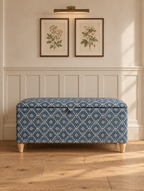 A blue and white patterned storage bench with wooden legs, set against a white paneled wall with two framed botanical prints and a wall lamp above.