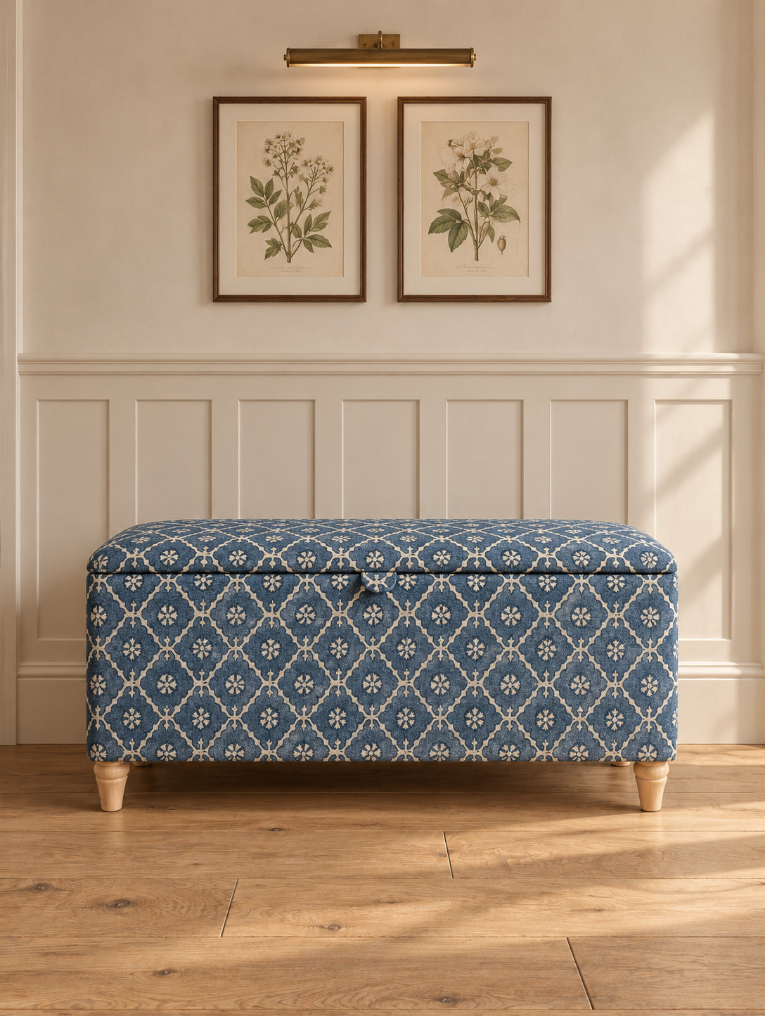 A blue and white patterned storage bench with wooden legs, set against a white paneled wall with two framed botanical prints and a wall lamp above.