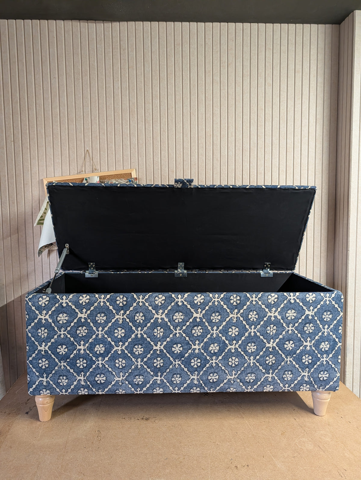 A fabric-covered storage bench with an open lid, featuring a blue and white floral trellis pattern and light wood legs, set against a beige paneled wall.
