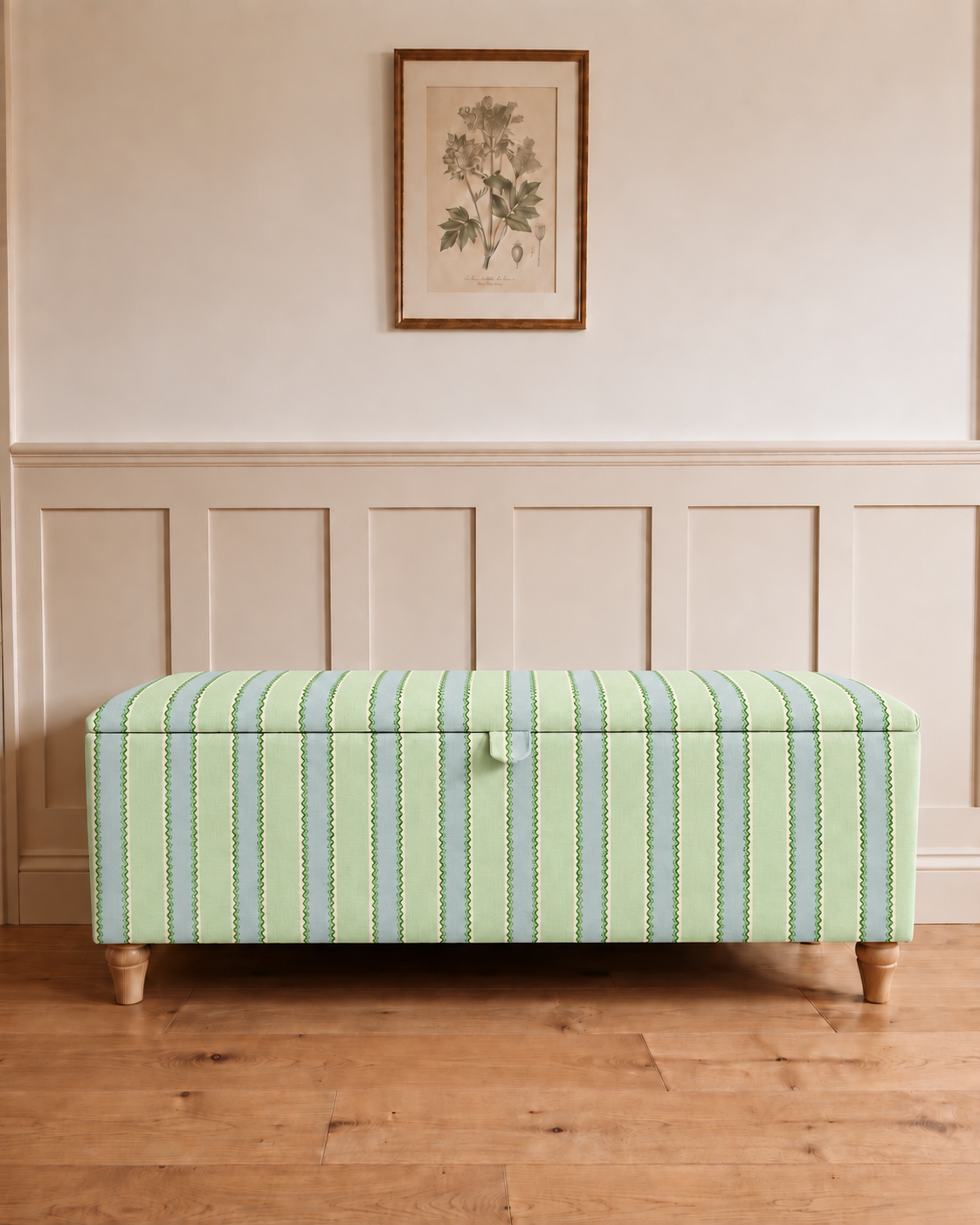 A storage bench upholstered in a light green and blue striped fabric with wooden legs, set against a cream paneled wall with a framed botanical print above.