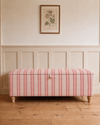 A rectangular storage bench upholstered in pink and white vertically striped fabric with light wood legs, placed against a cream paneled wall with a framed botanical print above.