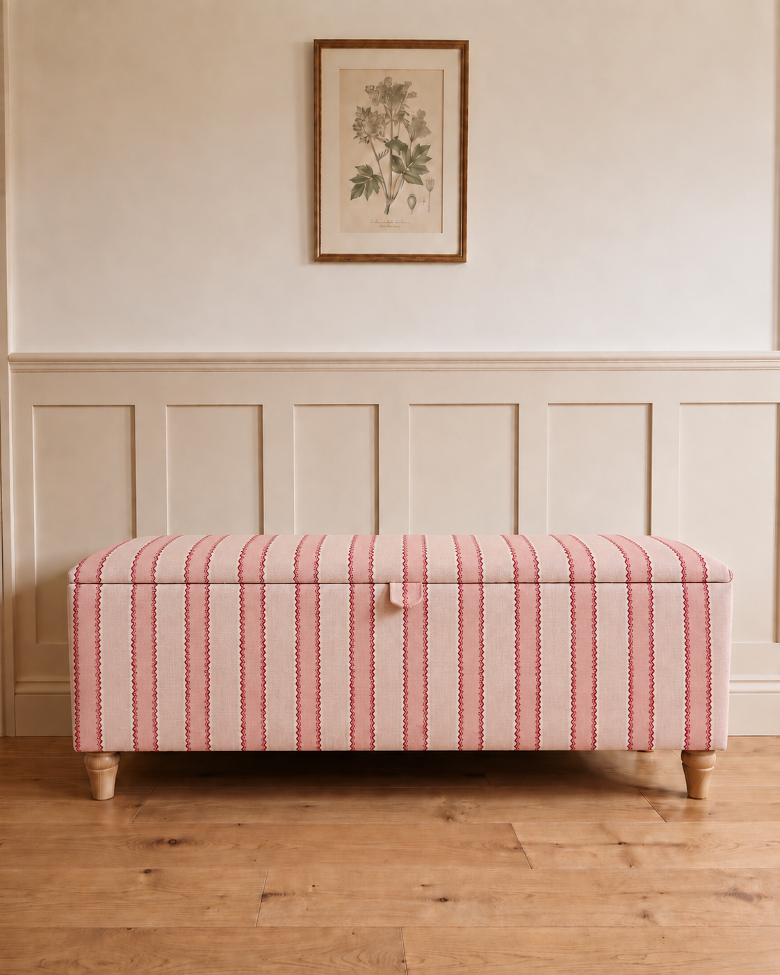 A rectangular storage bench upholstered in pink and white vertically striped fabric with light wood legs, placed against a cream paneled wall with a framed botanical print above.