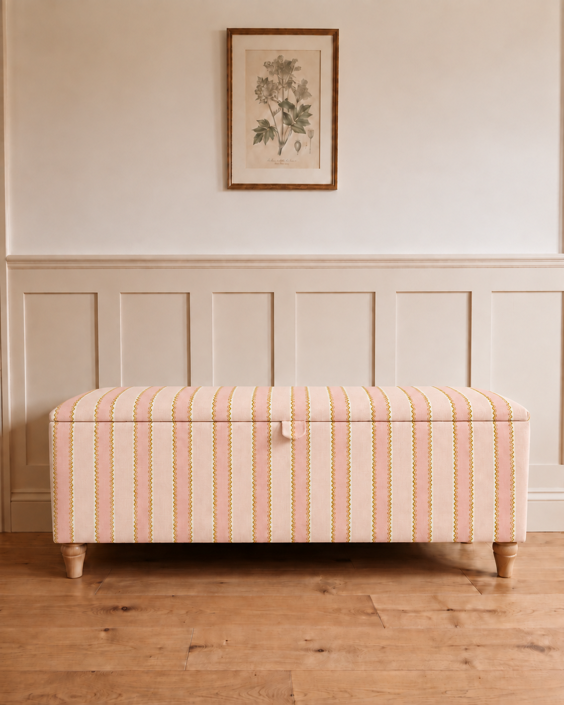 A rectangular upholstered storage bench with a cream base and vertical stripes in pink, orange, and yellow, set against a wainscoted wall with a framed botanical print above.