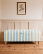 A square upholstered storage bench with a cream and light blue striped pattern, supported by four turned wooden legs, set against a classic wainscoted wall with a framed botanical print.