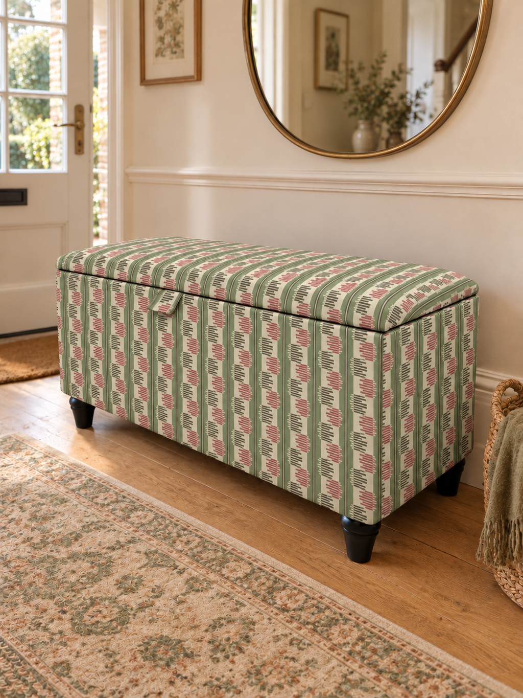 An upholstered storage bench with a green, pink, and white striped patterned fabric, set in a bright entryway on a wooden floor next to a patterned rug.
